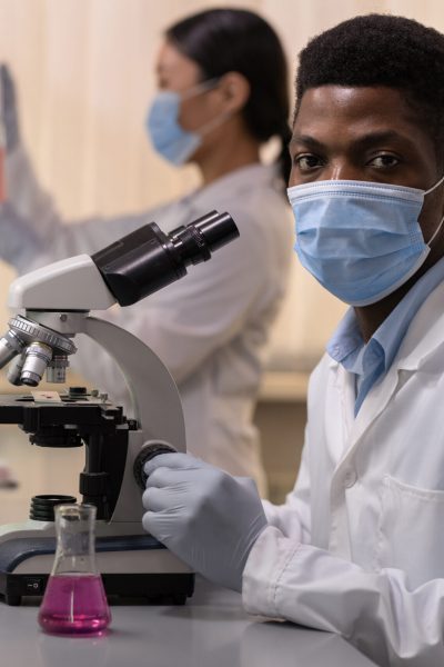 Portrait of African young chemist in mask looking at camera while working with microscope with his colleagues in the lab